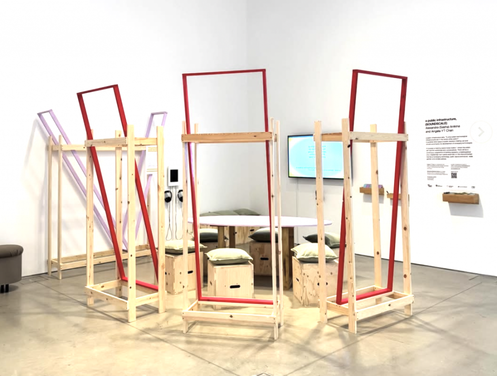 an art installation in a gallery space composed of wooden frames assembled in a loose circle around a wooden table. 4 frames are painted red and pink, the rest are untreated plain wood colour. 